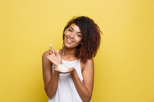 Beauty-Konzept - Schöne junge afroamerikanische Frau glücklich mit Hautpflege Creme. Gelb Studio Hintergrund. Text kopieren