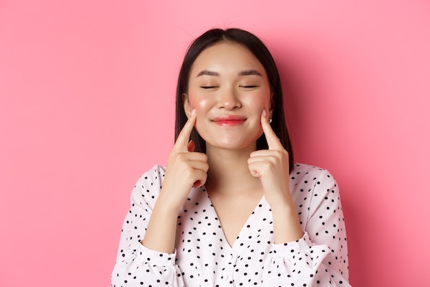 Beauty- und Lifestyle-Konzept. Nahaufnahme einer schönen asiatischen Frau, die mit geschlossenen Augen in die Wangen stößt, zufrieden lächelt und über rosafarbenem Hintergrund steht.