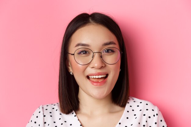 Beauty- und Lifestyle-Konzept. Nahaufnahme eines süßen asiatischen weiblichen Modells mit trendiger Brille, das glücklich in die Kamera lächelt und auf rosafarbenem Hintergrund steht.