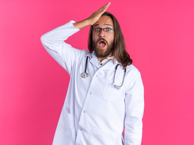 Bedauern eines erwachsenen männlichen Arztes, der ein medizinisches Gewand und ein Stethoskop mit Brille trägt und in die Kamera schaut, die Hand auf dem Kopf isoliert auf rosa Wand hält