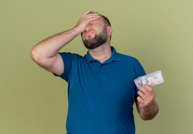 Bedauern erwachsenen erwachsenen slawischen Mann, der Geld hält und Hand auf die Stirn mit geschlossenen Augen legt