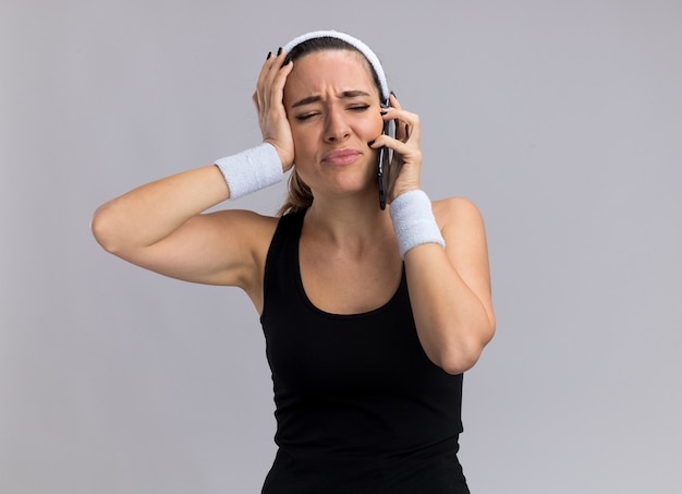 Bedauern junges hübsches sportliches mädchen mit stirnband und armbändern, das am telefon spricht und auf die seite schaut, die hand auf dem kopf isoliert auf weißer wand mit kopierraum