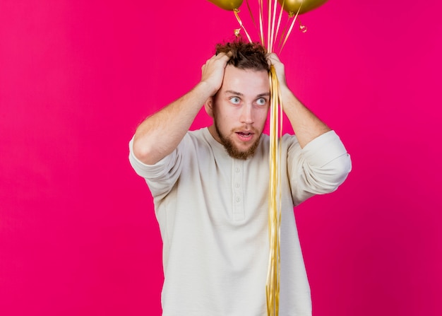 Bedauernder junger hübscher slawischer Partei-Typ, der Luftballons hält, die Hände auf Kopf halten, die gerade lokal auf purpurrotem Hintergrund mit Kopienraum suchen