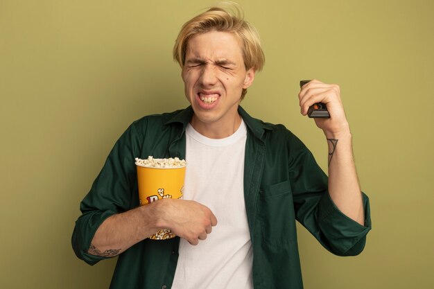 Bedauert mit geschlossenen Augen junger blonder Kerl, der grünes T-Shirt hält Eimer Popcorn hält und TV-Fernbedienung hebt