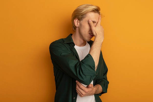 Bedauerter junger blonder Kerl, der grünes T-Shirt bedecktes Gesicht mit Hand trägt