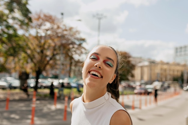 beeindruckendes mädchen mit hellem urlaubs-make-up lacht draußen im sonnenlicht mit einem glücklichen lächeln auf stadthintergrund