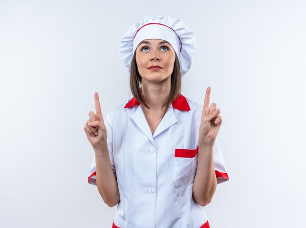 Beeindruckt, die junge Köchin mit Kochuniform nach oben zeigt, die auf weißer Wand isoliert ist?