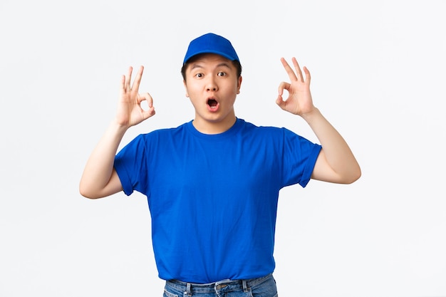 Beeindruckt und erstaunt reagieren asiatische Kurier in blauer Uniform auf super cooles Promo-Angebot. Lieferbote in Mütze und T-Shirt zeigt eine gute Geste, lobt gute Arbeit, gut gemacht, Kompliment tolle Wahl