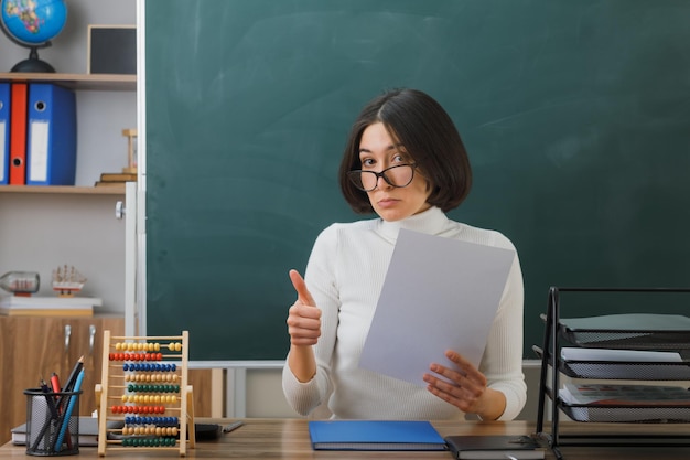 beeindruckt zeigt Daumen nach oben Junge Lehrerin mit Brille, die Papier hält und am Schreibtisch mit Schulwerkzeugen im Klassenzimmer sitzt