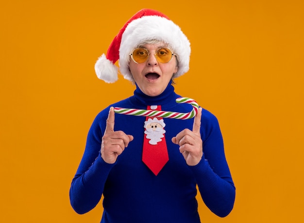 Beeindruckte ältere Frau in Sonnenbrille mit Weihnachtsmütze und Weihnachtsmann-Krawatte mit Zuckerstange isoliert auf orangefarbener Wand mit Kopierraum