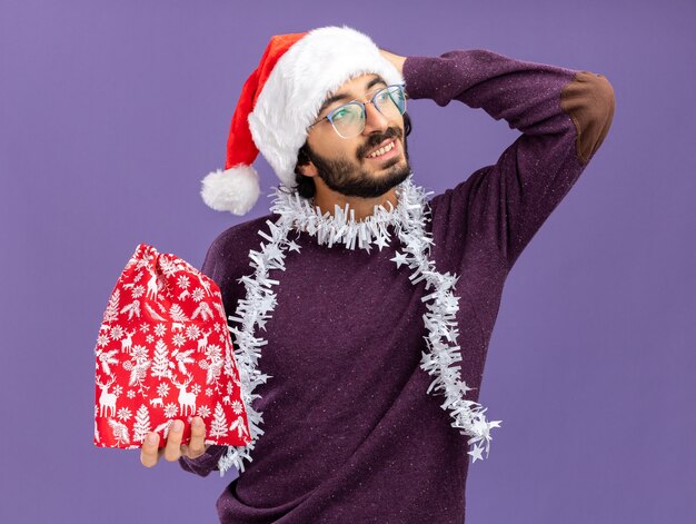 Beeindruckte aussehende Seite junger hübscher Kerl, der Weihnachtsmütze mit Girlande am Hals hält, der Weihnachtstasche hält Hand hinter Kopf lokalisiert auf blauer Wand