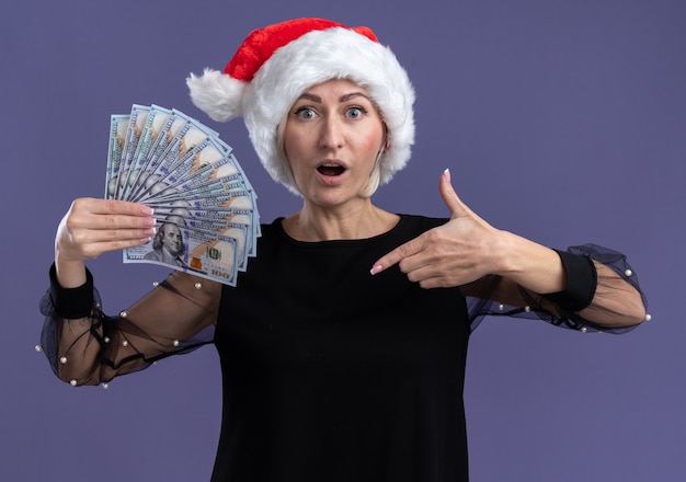 Beeindruckte blonde Frau mittleren Alters, die Weihnachtshut hält und auf Geld zeigt, das auf lila Wand lokalisiert ist