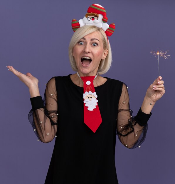 Beeindruckte blonde Frau mittleren Alters, die Weihnachtsmann-Stirnband und Krawatte hält, die Feiertagswunderkerze hält, die Kamera zeigt, die leere Hand lokalisiert auf lila Hintergrund zeigt