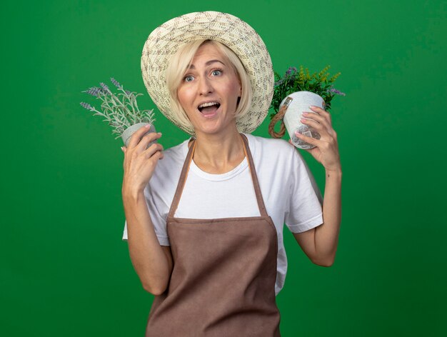 Beeindruckte blonde Gärtnerin mittleren Alters in Uniform mit Hut, die Blumentöpfe in der Nähe des Kopfes hält und auf die Seite isoliert auf grüner Wand schaut