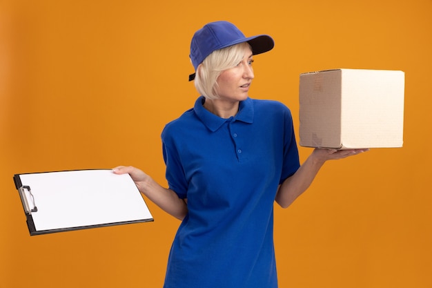 Beeindruckte blonde Lieferfrau mittleren Alters in blauer Uniform und Mütze, die Karton und Zwischenablage mit Blick auf die Seite hält