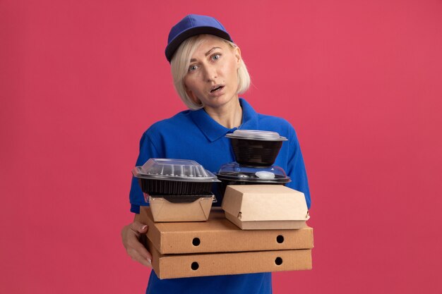 Beeindruckte blonde Lieferfrau mittleren Alters in blauer Uniform und Mütze, die Pizzapakete mit Lebensmittelbehältern und Papiernahrungspaketen auf ihnen hält, die nach vorne schauen