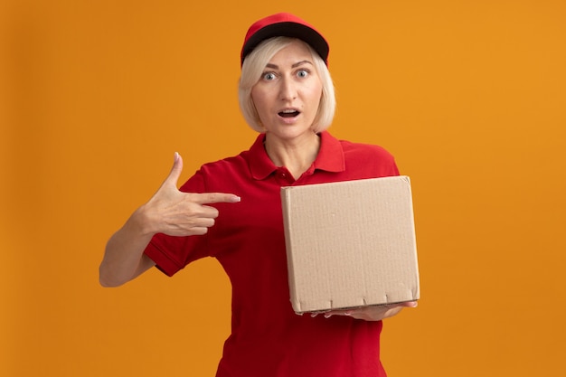 Beeindruckte blonde Lieferfrau mittleren Alters in roter Uniform und Mütze, die auf Karton hält und auf die Vorderseite zeigt, die auf orangefarbener Wand mit Kopierraum isoliert ist point