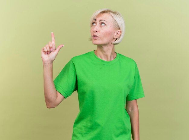 Beeindruckte blonde slawische Frau mittleren Alters, die den Kopf zur Seite dreht und lokal auf olivgrüne Wand mit Kopienraum zeigt