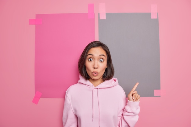 Beeindruckte emotionale junge asiatin keucht von schockpunkten mit dem finger nach oben zeigt etwas unerwartetes banner nach oben gekleidet in hoodie-posen gegen rosa wand schlägt sonderverkauf vor