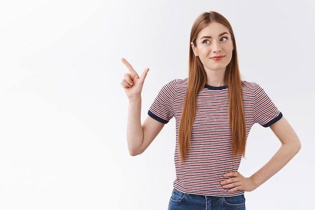 Beeindruckte, enthusiastische, gut aussehende kaukasische Frau in gestreiftem T-Shirt, schräger Kopf und amüsiertes Grinsen, zeigend auf die obere linke Ecke, schauen Sie sich etwas Interessantes und Cooles, weißen Hintergrund an