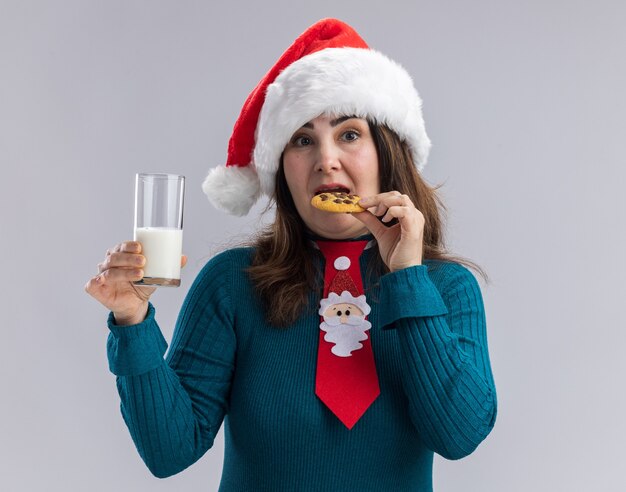 Beeindruckte erwachsene kaukasische Frau mit Weihnachtsmütze und Weihnachtsmann-Krawatte, die ein Glas Milch hält und Plätzchen isst, isoliert auf weißer Wand mit Kopierraum