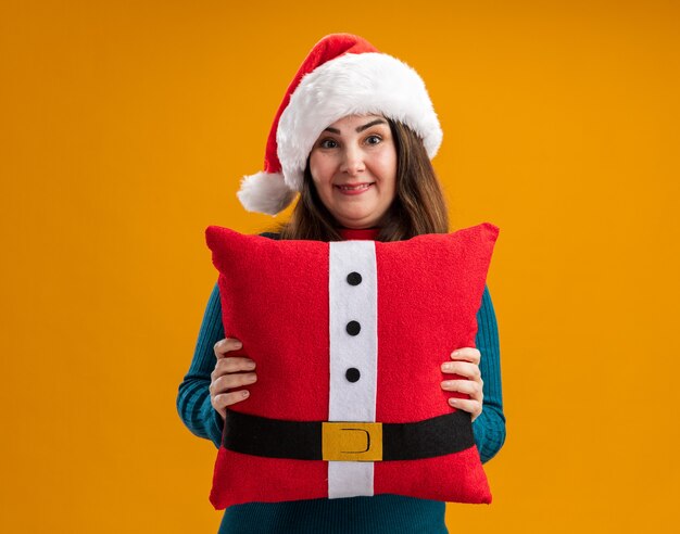 Beeindruckte erwachsene kaukasische Frau mit Weihnachtsmütze und Weihnachtsmannkrawatte, die dekoriertes Kissen isoliert auf oranger Wand mit Kopierraum hält