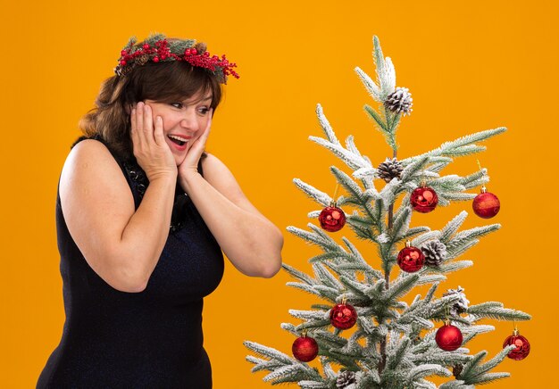 Beeindruckte Frau mittleren Alters, die Weihnachtskopfkranz und Lametta-Girlande um den Hals trägt, die in der Profilansicht nahe geschmücktem Weihnachtsbaum stehen