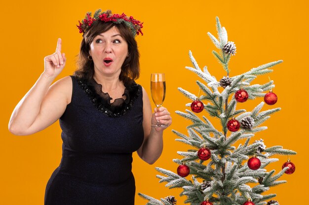 Beeindruckte Frau mittleren Alters, die Weihnachtskopfkranz und Lametta-Girlande um den Hals trägt, die nahe geschmücktem Weihnachtsbaum hält, der Glas Champagner hält