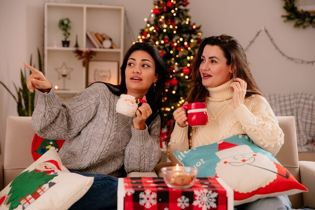 Beeindruckte hübsche junge Mädchen halten Tassen und schauen zur Seite, sitzen auf Sesseln und genießen die Weihnachtszeit zu Hause
