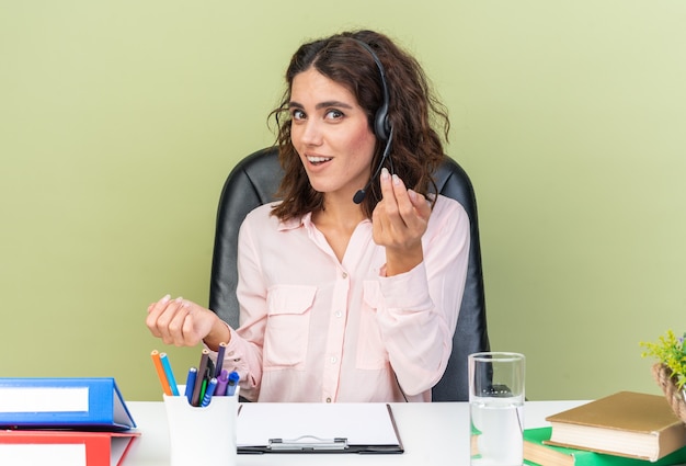 Beeindruckte hübsche kaukasische Callcenter-Betreiberin auf Kopfhörern, die am Schreibtisch mit Bürowerkzeugen sitzen und ihre Hände offen halten