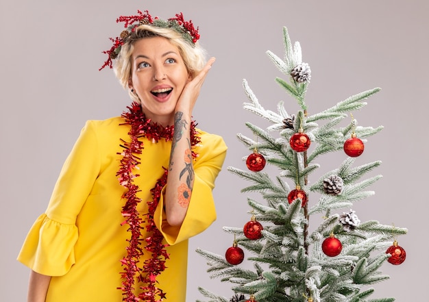 Beeindruckte junge blonde Frau, die den Weihnachtskopfkranz und die Lametta-Girlande um den Hals trägt, die nahe beim verzierten Weihnachtsbaum steht, der Gesicht berührt, das Seite lokalisiert auf weißem Hintergrund betrachtet