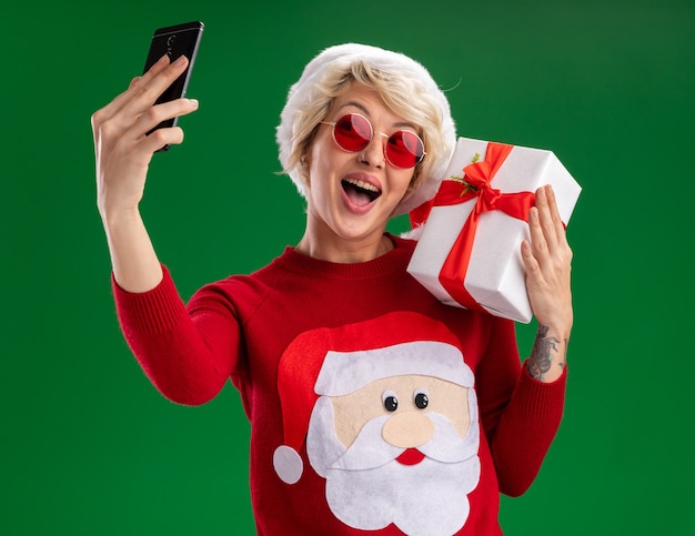 Beeindruckte junge blonde frau, die weihnachtsmütze und weihnachtsmannpullover des weihnachtsmannes mit brille hält, die geschenkpaket auf schulter nimmt, das selfie lokalisiert auf grünem hintergrund nimmt