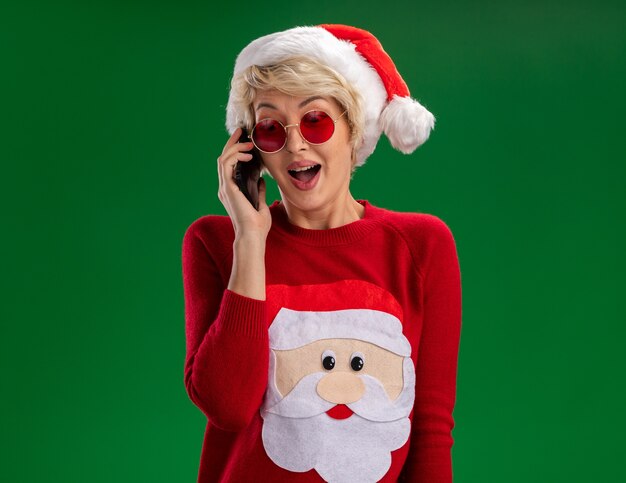 Beeindruckte junge blonde Frau, die Weihnachtsmütze und Weihnachtsmannpullover des Weihnachtsmannes mit Brille trägt, die am Telefon sprechen, das lokalisiert auf grünem Hintergrund schaut