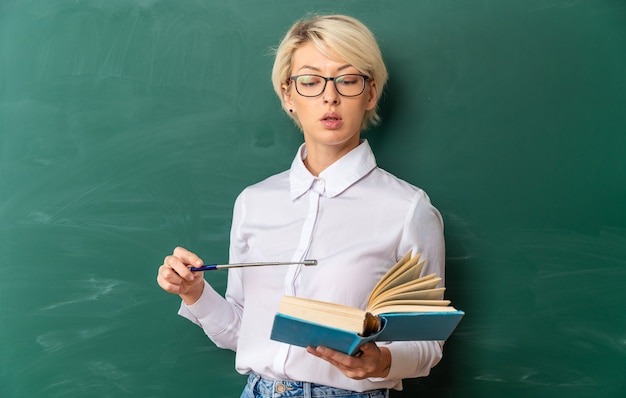 Beeindruckte junge blonde Lehrerin, die eine Brille im Klassenzimmer trägt, die vor einer Tafel steht, die liest und mit einem Zeiger auf ein Buch mit Kopienraum zeigt