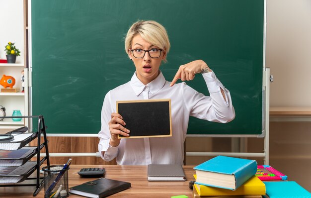 Beeindruckte junge blonde Lehrerin mit Brille, die am Schreibtisch mit Schulmaterial im Klassenzimmer sitzt und auf die Mini-Tafel mit Blick auf die Vorderseite zeigt und zeigt