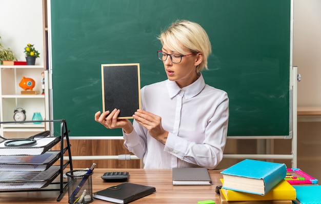 Kostenloses Foto beeindruckte junge blonde lehrerin mit brille, die am schreibtisch mit schulmaterial im klassenzimmer sitzt und eine mini-tafel zeigt, die sie anschaut