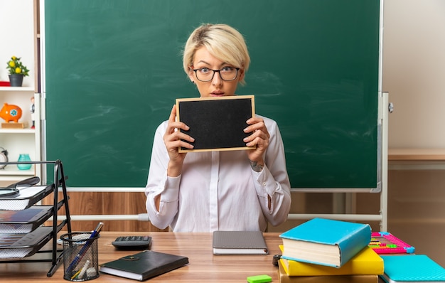 Beeindruckte junge blonde Lehrerin mit Brille, die am Schreibtisch mit Schulwerkzeugen im Klassenzimmer sitzt und eine Mini-Tafel mit Blick auf die Kamera zeigt