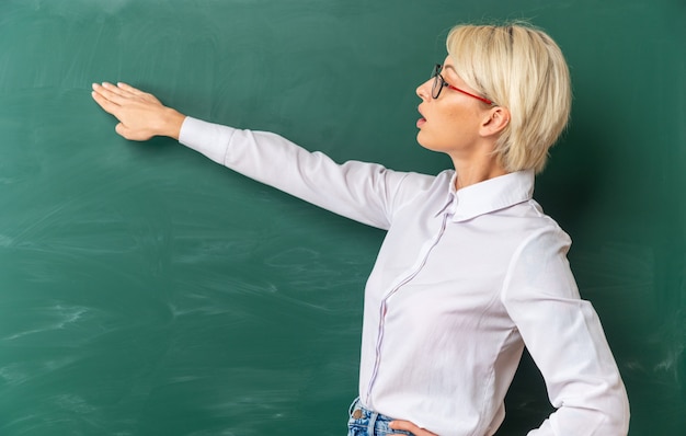 Beeindruckte junge blonde Lehrerin mit Brille im Klassenzimmer, die in der Profilansicht vor der Tafel steht und auf die Tafel mit der Hand zeigt, die eine andere Hand auf der Taille hält