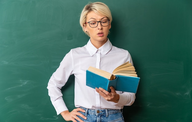 Beeindruckte junge blonde Lehrerin mit Brille im Klassenzimmer, die vor der Tafel steht, die ein Buch hält und liest und die Hand auf der Taille hält