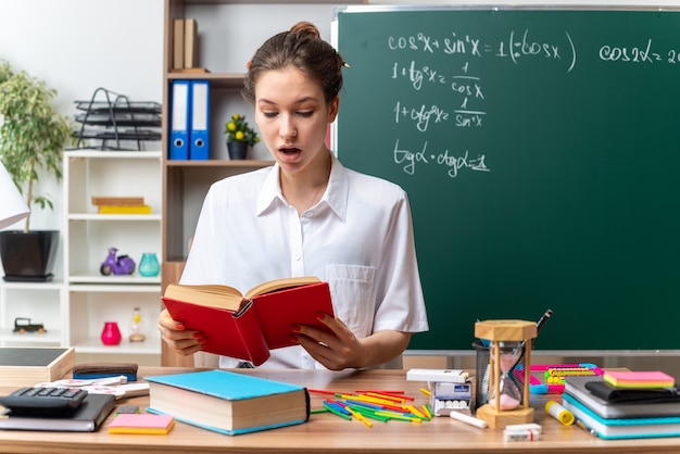 Beeindruckte junge blonde Mathematiklehrerin sitzt am Schreibtisch mit Schulwerkzeugen und liest Buch im Klassenzimmer