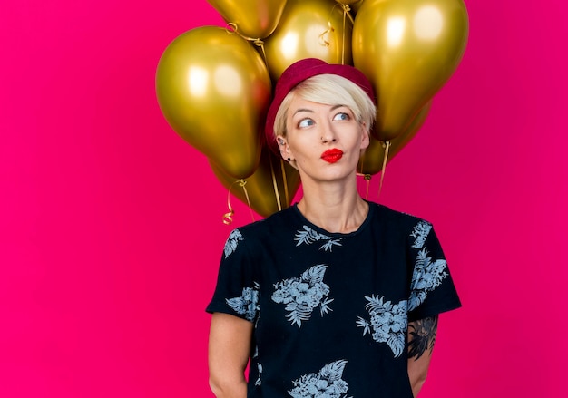 Beeindruckte junge blonde Partyfrau, die Partyhut trägt, der vor Ballons steht, die Seite betrachten, die Hände hinter dem Rücken lokalisiert auf rosa Wand mit Kopienraum hält