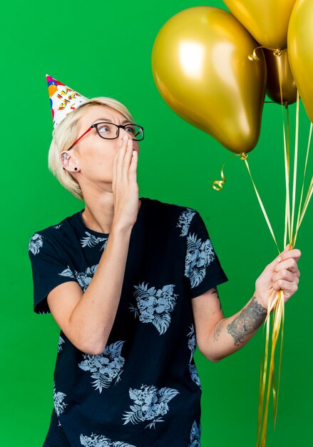 Beeindruckte junge blonde Partygirl, die Brille und Geburtstagskappe hält und Ballons flüstert, die lokalisiert auf grünem Hintergrund halten