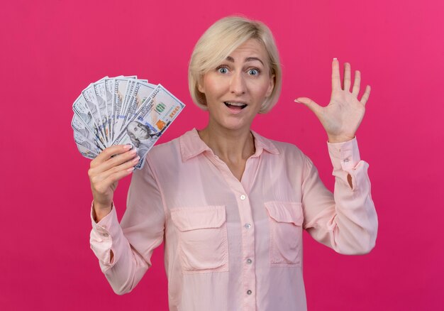 Beeindruckte junge blonde slawische Frau, die Geld hält und Hallo mit Hand lokalisiert auf rosa Hintergrund gestikuliert