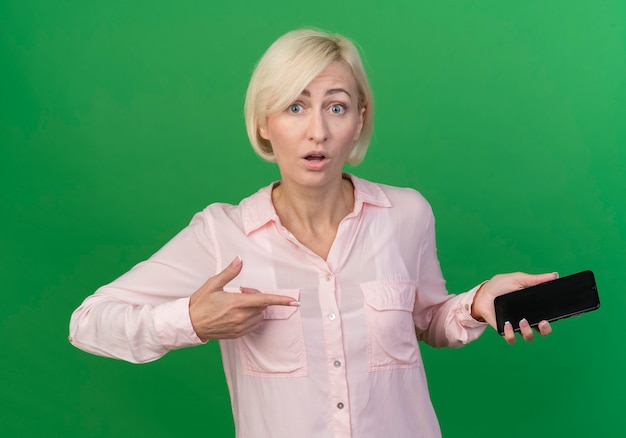 Beeindruckte junge blonde slawische Frau hält und zeigt auf Handy lokalisiert auf grünem Hintergrund