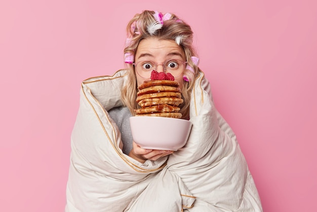 Beeindruckte junge europäerin mit haarrollen, die in eine bettdecke gehüllt sind, hält eine schüssel mit appetitlichen pfannkuchen mit sirup und trägt eine große brille, die über rosafarbenem hintergrund isoliert ist. konzept für frühstück und morgenzeit