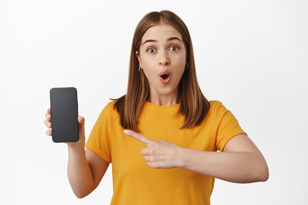Beeindruckte junge Frau, die mit dem Finger auf den Verkauf des Smartphone-Bildschirms zeigt, erstaunt nach Luft schnappt und ein tolles Promo-Angebot auf dem Handy zeigt, weiße Wand.