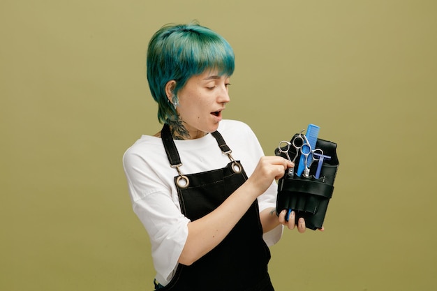 Beeindruckte junge Friseurin in Uniform, die eine Friseurtasche mit Friseurwerkzeugen hält, die eine Schere herausziehen, isoliert auf olivgrünem Hintergrund