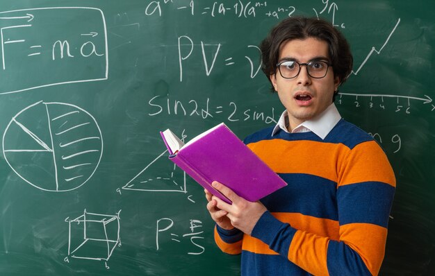 Beeindruckte junge Geometrielehrerin mit Brille, die in der Profilansicht vor der Tafel im Klassenzimmer steht und ein offenes Buch mit Blick auf die Vorderseite hält