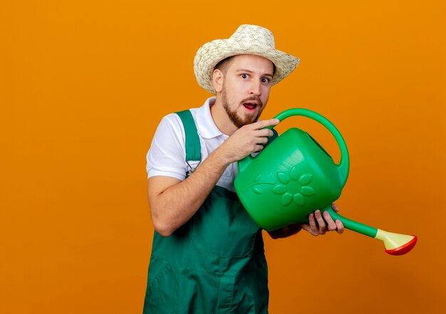 Beeindruckte junge gutaussehende slawische Gärtnerin in Uniform und Hut, die Bewässerung hält, die vorgibt, Bewässerung isoliert vorzutäuschen