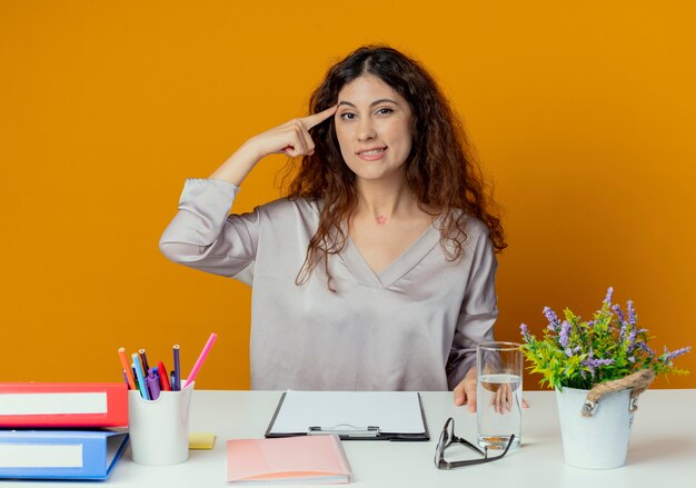 Beeindruckte junge hübsche Büroangestellte, die am Schreibtisch mit Bürowerkzeugen sitzt, die Finger auf Stirn lokalisiert auf orange Wand setzen